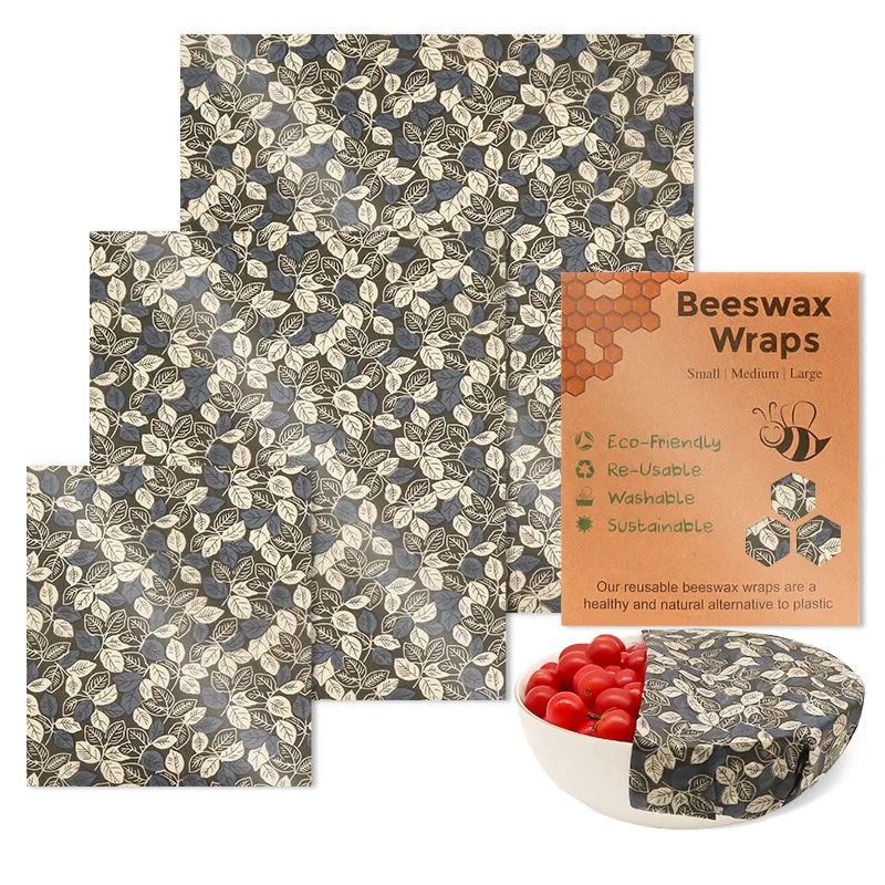 Three square reusable beeswax food wrap sheets in leaf pattern with packaging and a bowl of tomatoes.